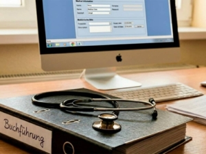 Digitale Buchführung Stethoskop auf einem Ordner mit der Aufschrift Buchführung vor einem Computerarbeitsplatz.